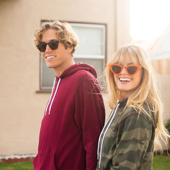 A man and woman, both in sunglasses, smile and are wearing a maroon hoodie and green camouflage hoodie, respectively.