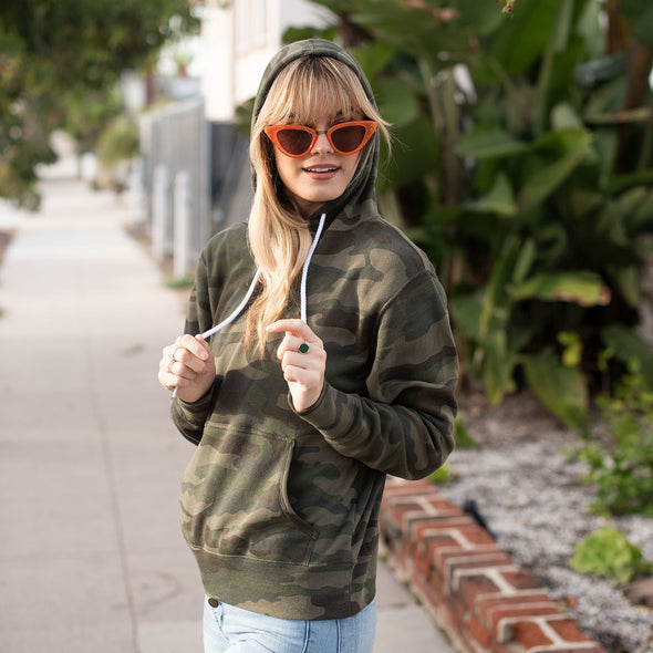 A woman poses in a green camouflage pullover hoodie with the hood on.