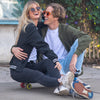 A woman and a man are laughing and sitting on a skateboard. The woman is wearing a black bomber jacket and the man is wearing an army green bomber jacket.