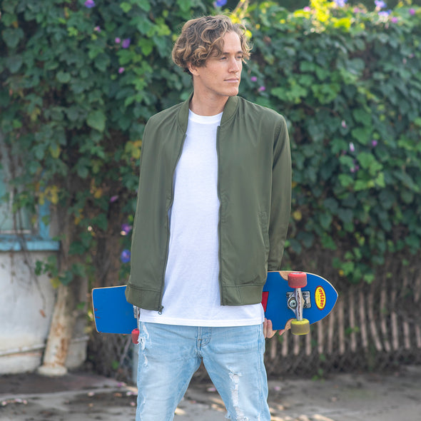 A man is holding a skateboard and wearing an army green bomber jacket over a white t-shirt.