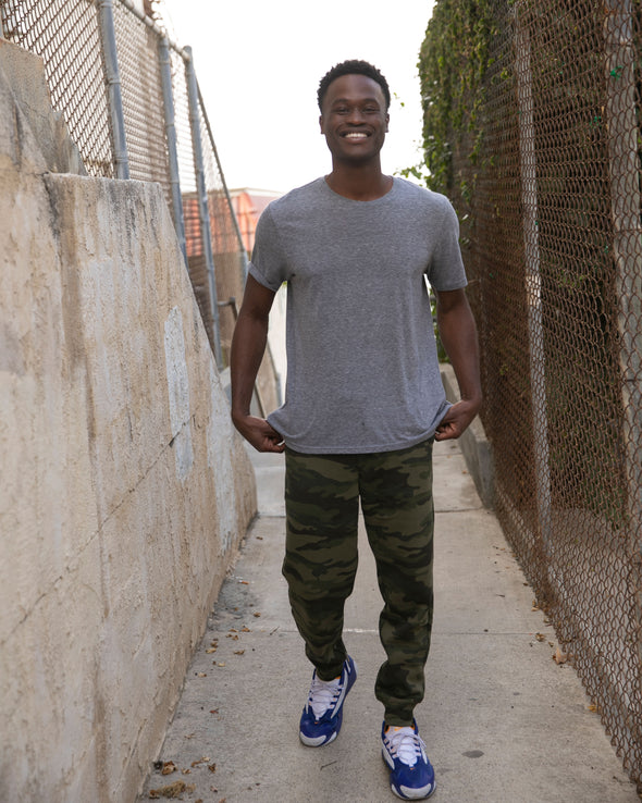 Man is posing in alley wearing a gray t-shirt and green camouflage sweatpants.