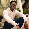 A man sits on a curb in an off-white crew neck sweater.