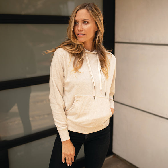 A woman poses in front of a building wearing a beige french terry pullover hoodie.