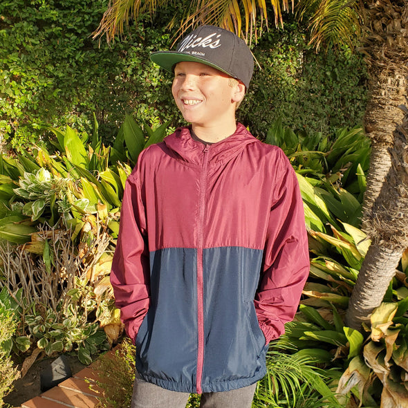 A boy is wearing a maroon and navy windbreaker hoodie in front of plants and foliage.