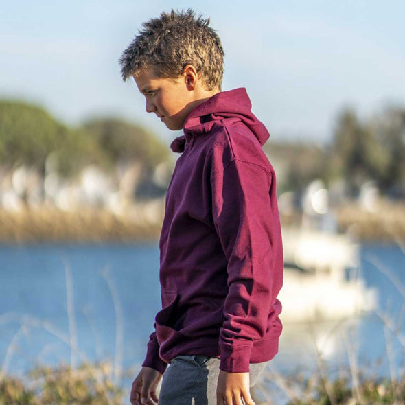 Boy walks in front of lake and he is wearing a maroon pullover hoodie.