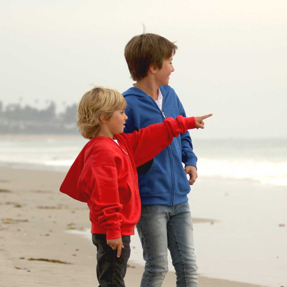 Two boys standing on the beach, looking out into the ocean. The boy on the right is wearing a blue hoodie and the boy on the left is wearing a red hoodie.