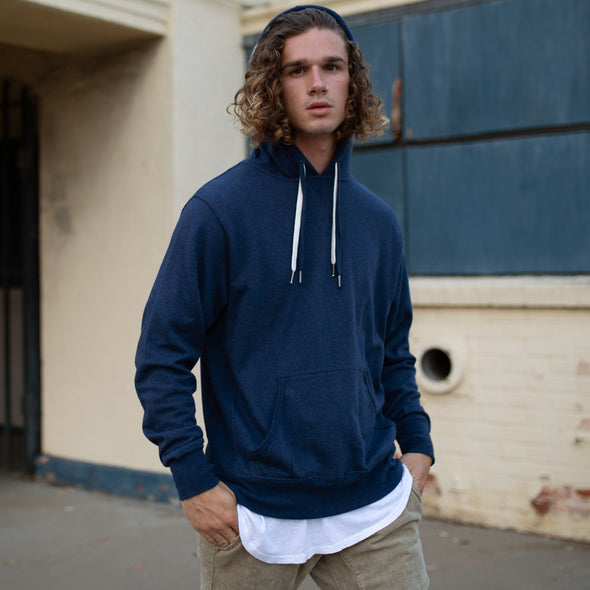 A man poses in front of a building wearing a navy pullover hoodie with a kangaroo pocket and two drawstrings.