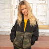 A girl is wearing a black and green camouflage zip-up windbreaker in front of an entrance building.