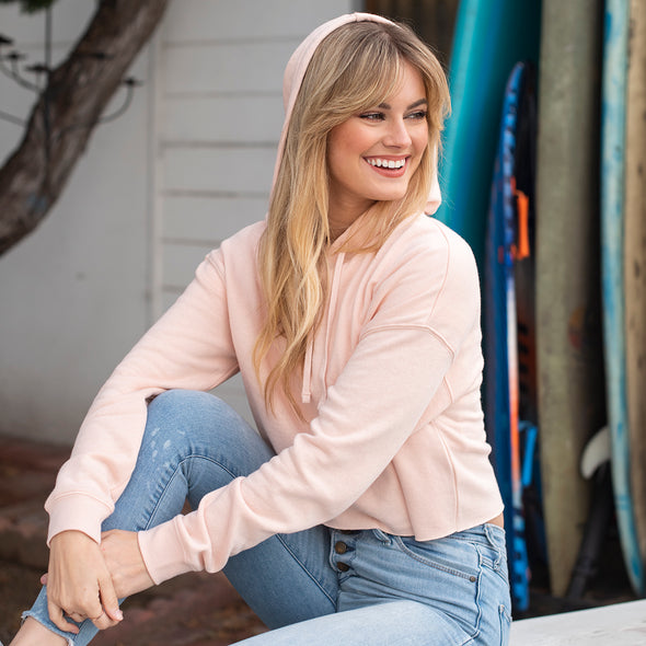 Woman poses on a bench in a pink crop top hoodie with the hood on.